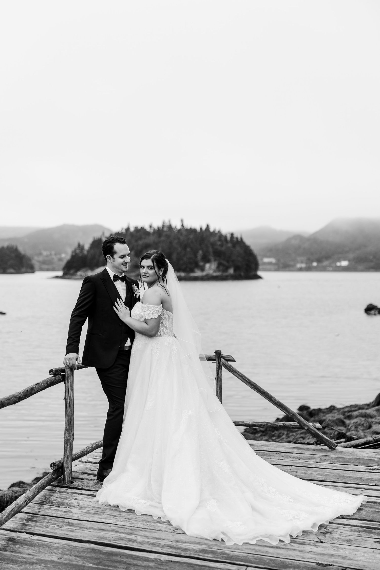 Black and white photo of bride and groom posing on wooden dock with island and mountains in background.
