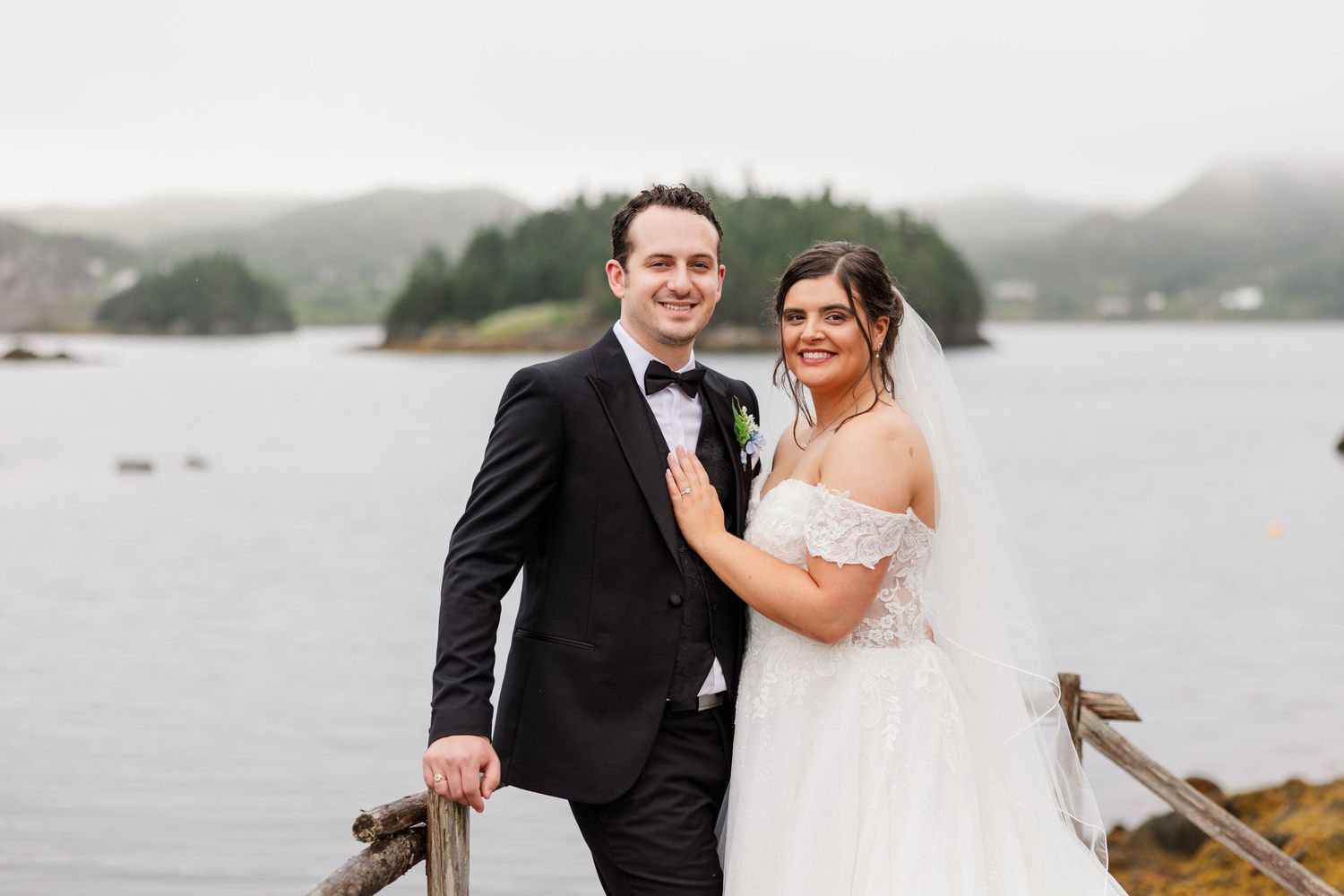 Bride and groom posing by a lakeside railing on a cloudy day, bride in off-shoulder gown, groom in black tuxedo.