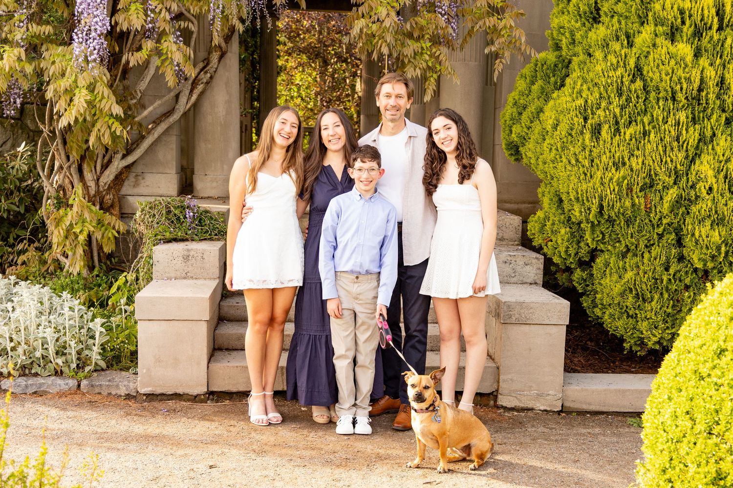 Family of five posing outdoors with a corgi dog, surrounded by lush greenery and stone steps in Connecticut during family photo session