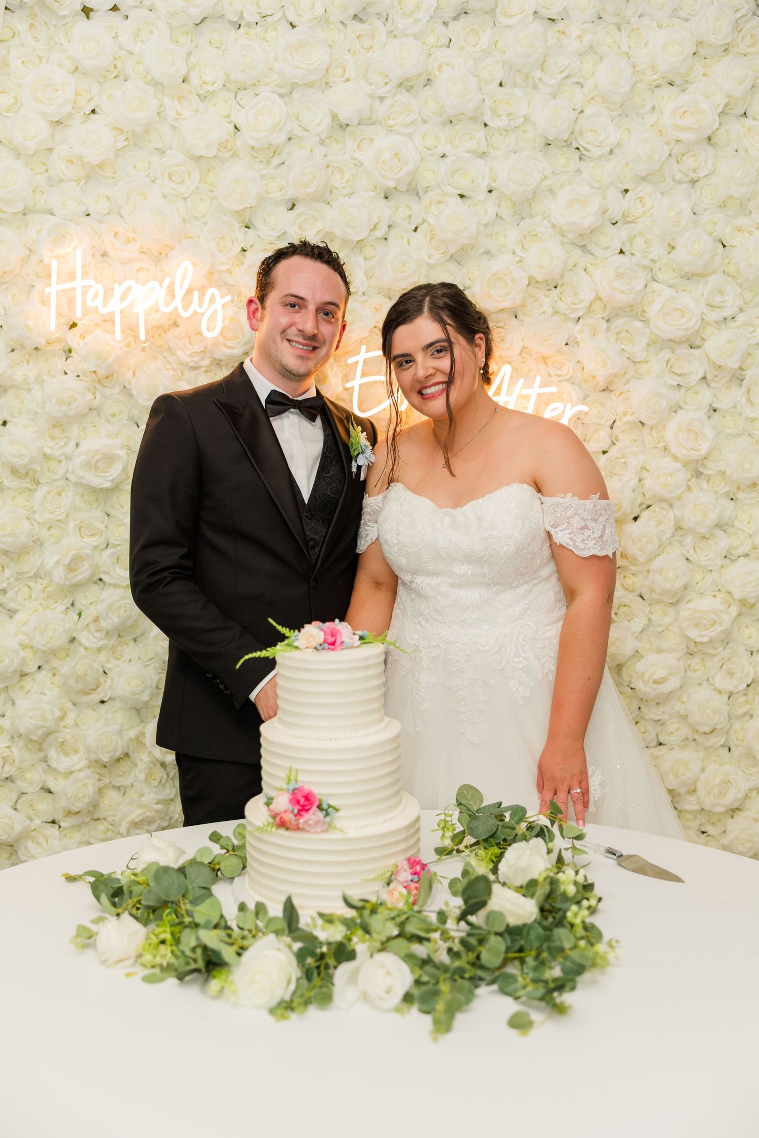 Newlyweds smiling beside a white tiered wedding cake in front of a floral wall with a Happily Ever After neon sign.