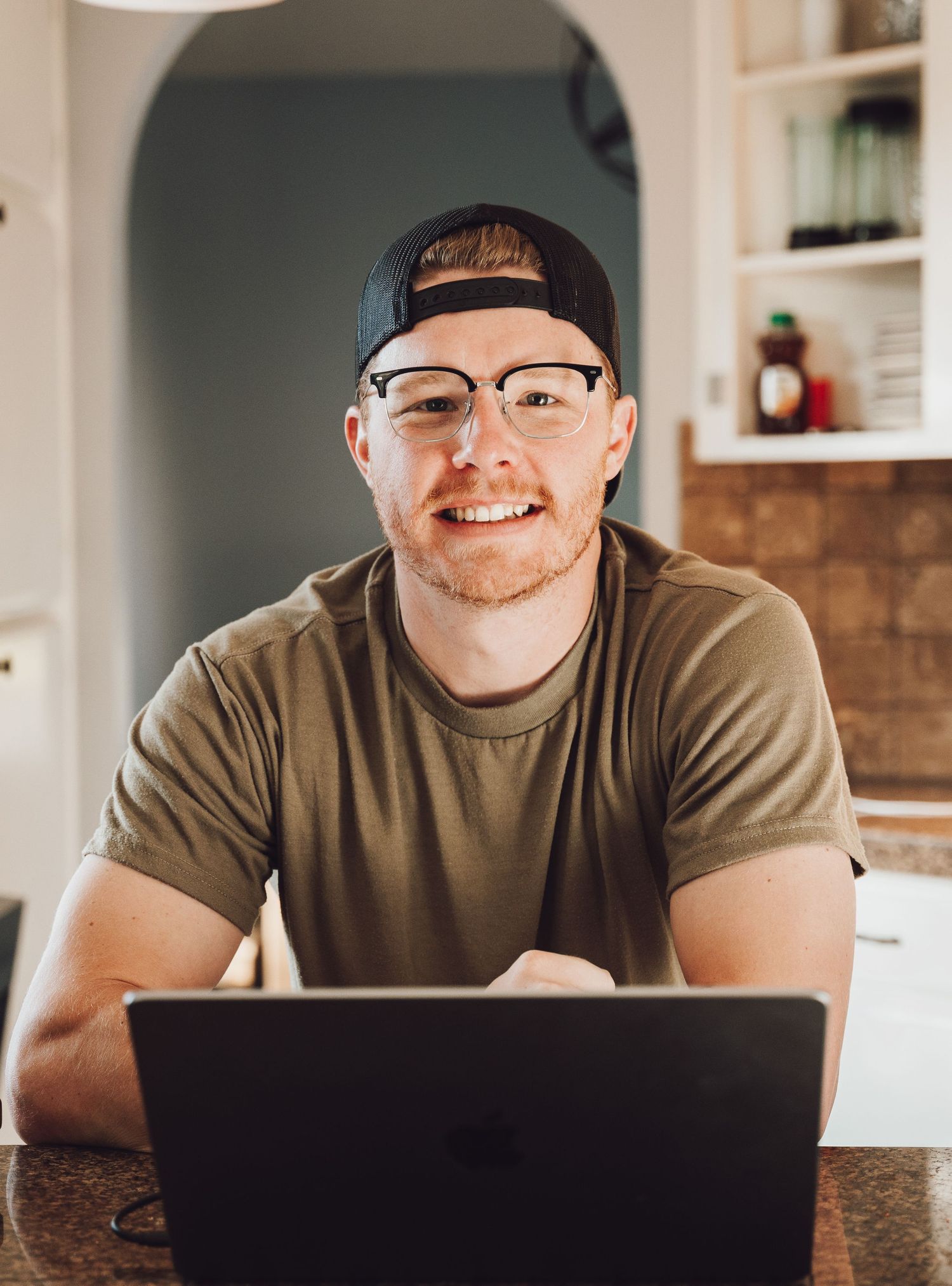Smiling young man with glasses and backward cap working on laptop at home desk in casual olive t-shirt.