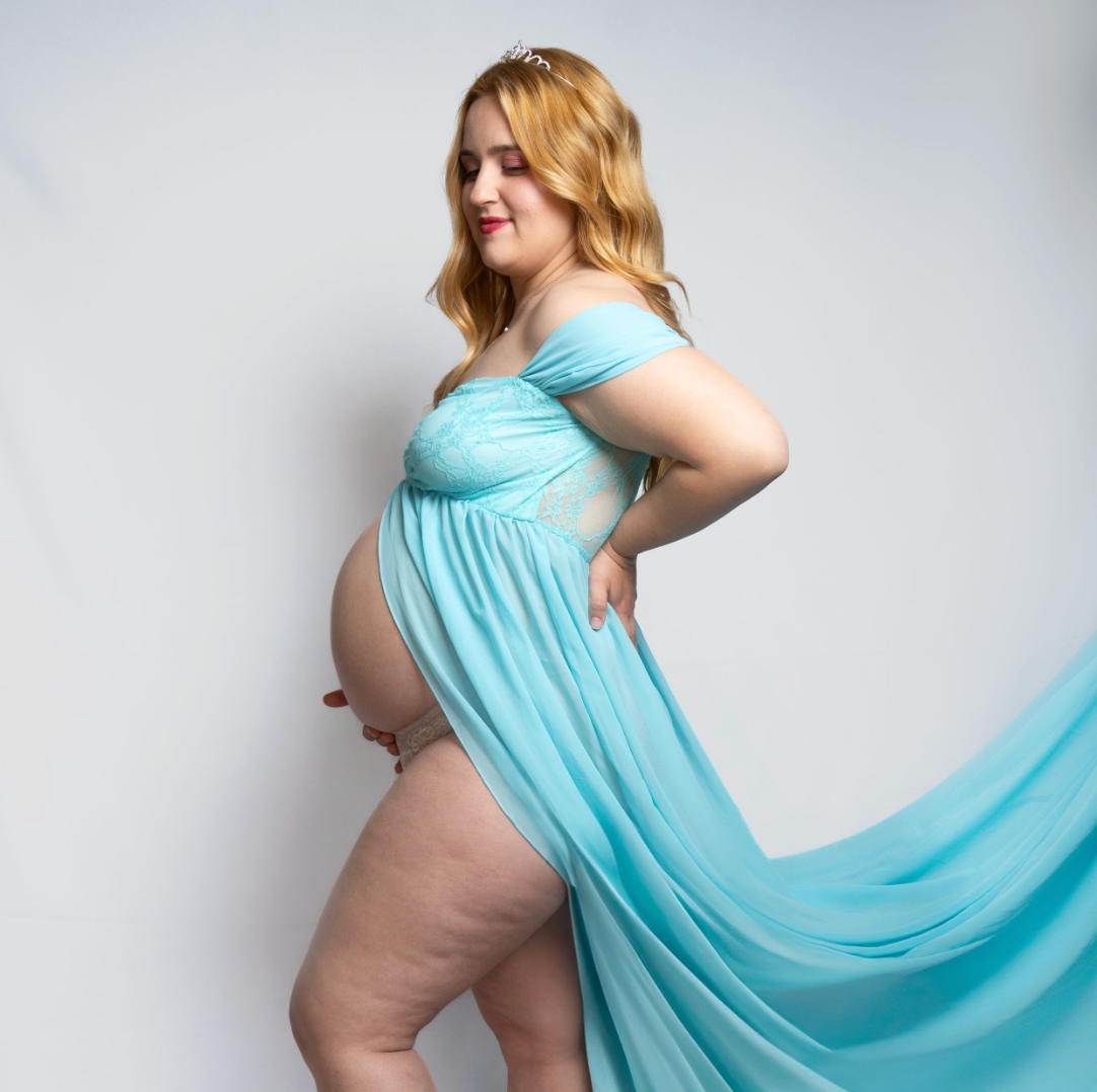 Pregnant woman in flowing teal maternity gown with off-shoulder design poses gracefully against white background.