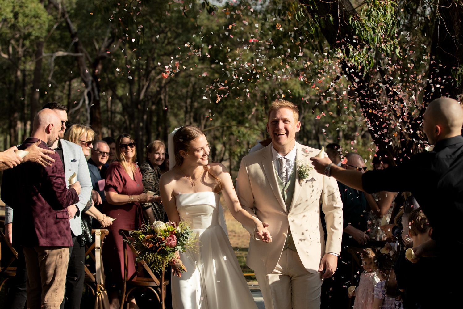A joyful bride and groom walk hand-in-hand through a confetti shower at their classic, elegant outdoor wedding ceremony.