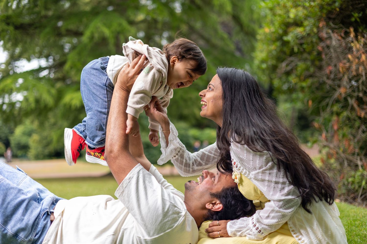 Happy family playing outdoors, father lifting baby while mother watches and smiles on green grass in a park.