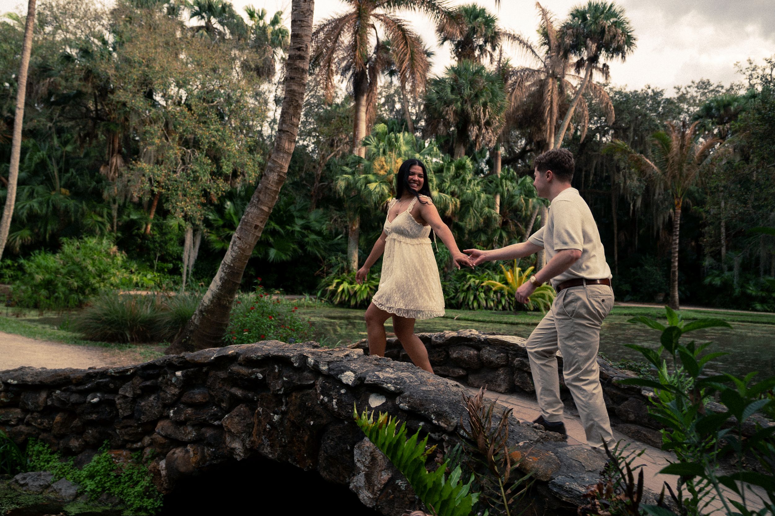 Couple holding hands on a stone bridge surrounded by lush tropical palm trees in a romantic outdoor setting.
