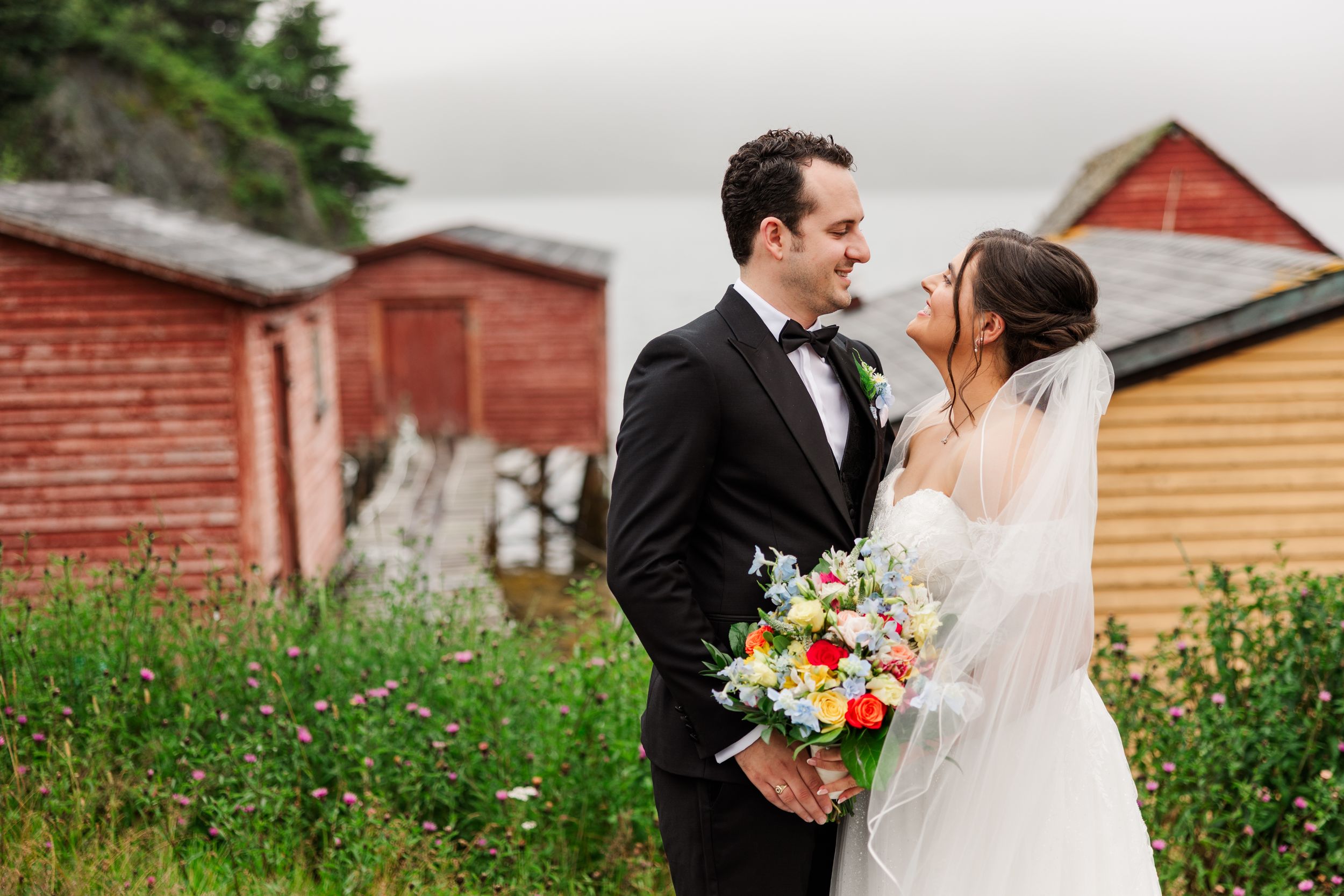 Bride and groom share a romantic moment outdoors near rustic red buildings, bride holding a colorful bouquet.