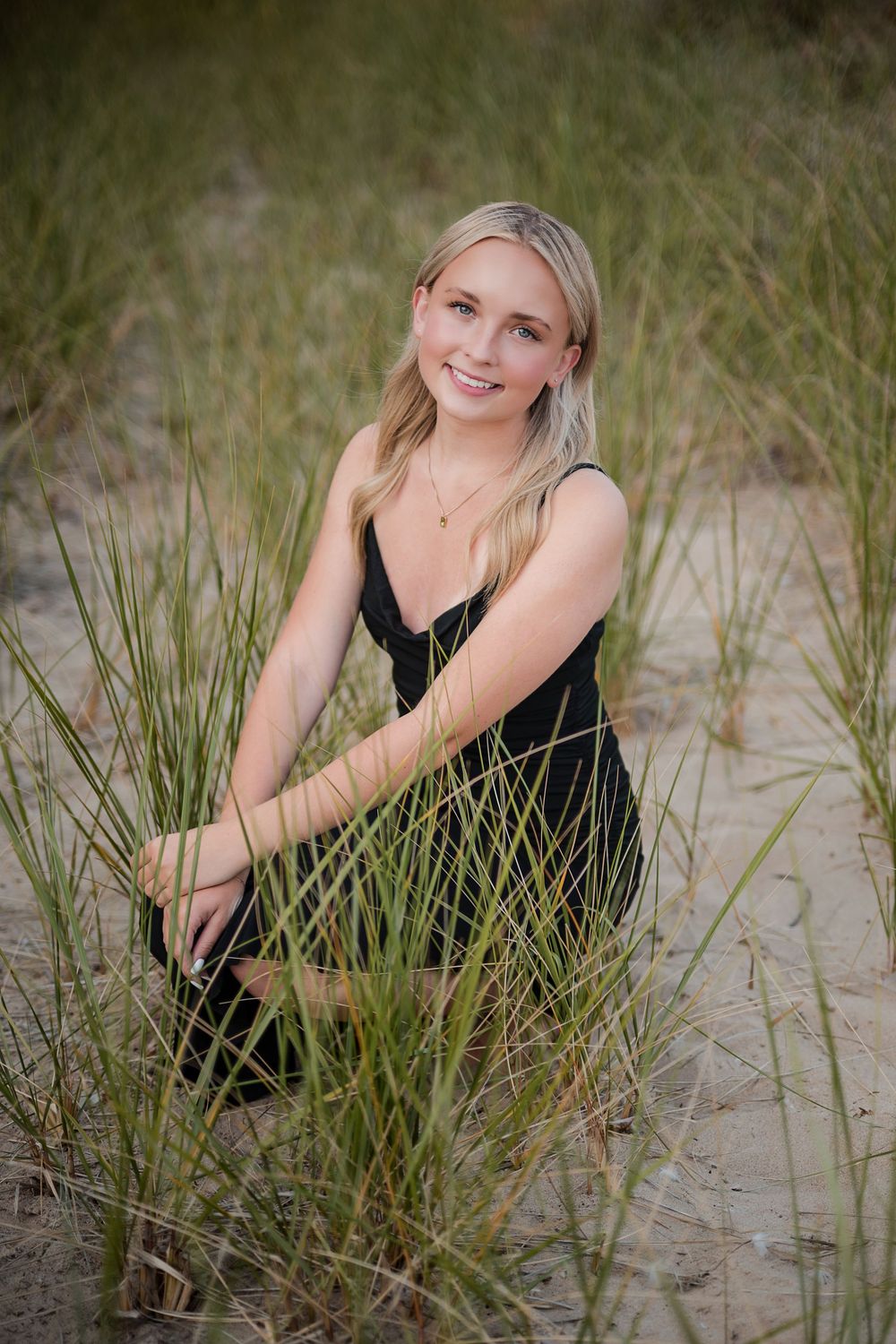 Young blonde woman in black dress sitting gracefully among beach grass on sandy dunes, smiling outdoors.