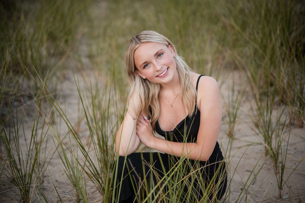 Smiling blonde woman in black dress posing among beach grass on sandy dunes in a senior portrait photo.