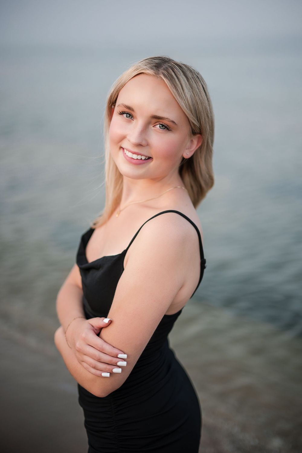 Young blonde woman in black dress smiling by the water's edge.