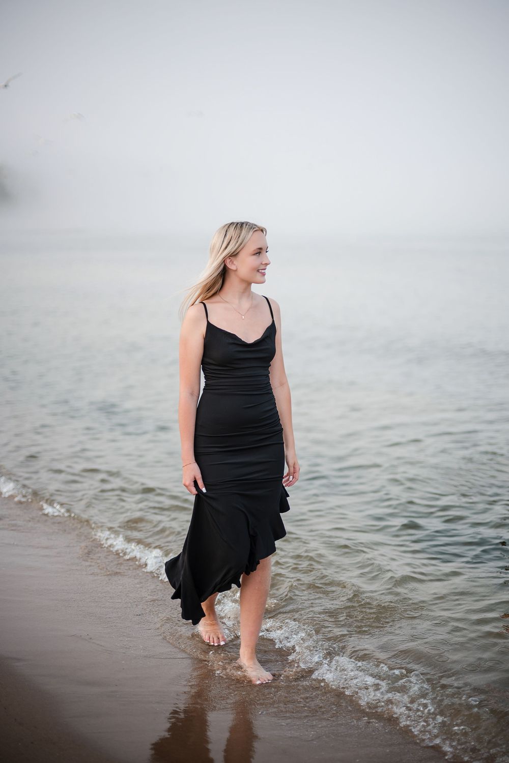 Smiling blonde woman in black ruffle dress walking barefoot along misty beach shoreline.