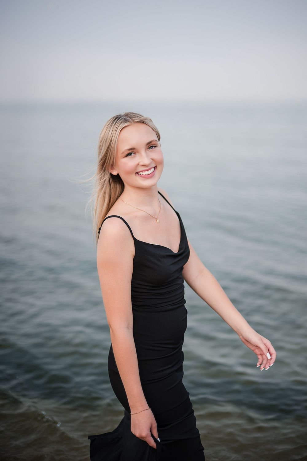 Smiling blonde woman in black spaghetti strap dress posing by the ocean on an overcast day.