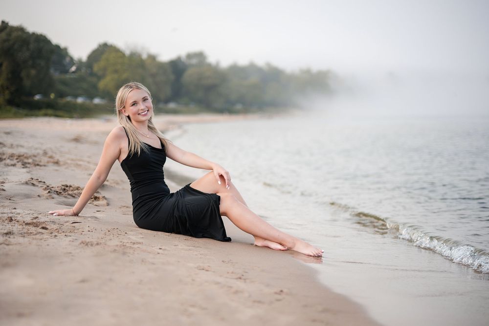 Smiling blonde woman in black dress sitting on sandy beach shore with misty trees and calm water in background.