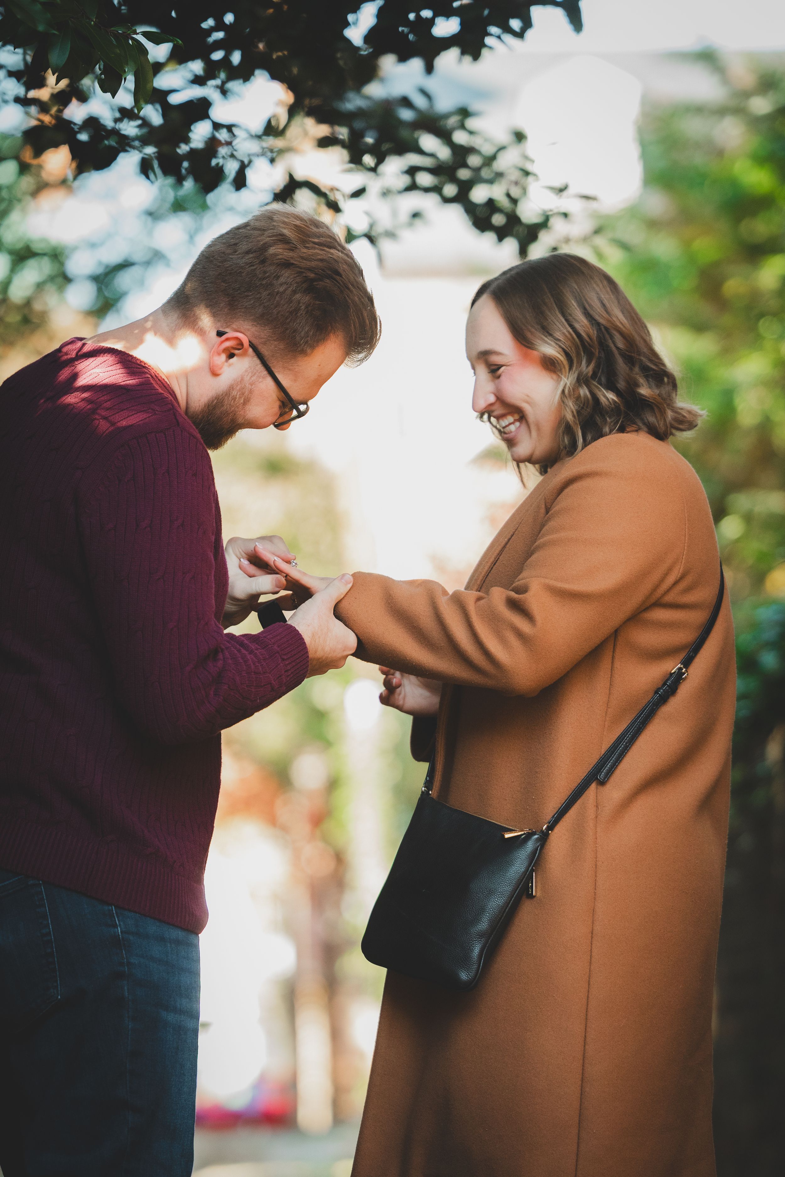 A man gently sliding an oval diamond engagement ring onto his bride's finger after proposing in a brick alleyway