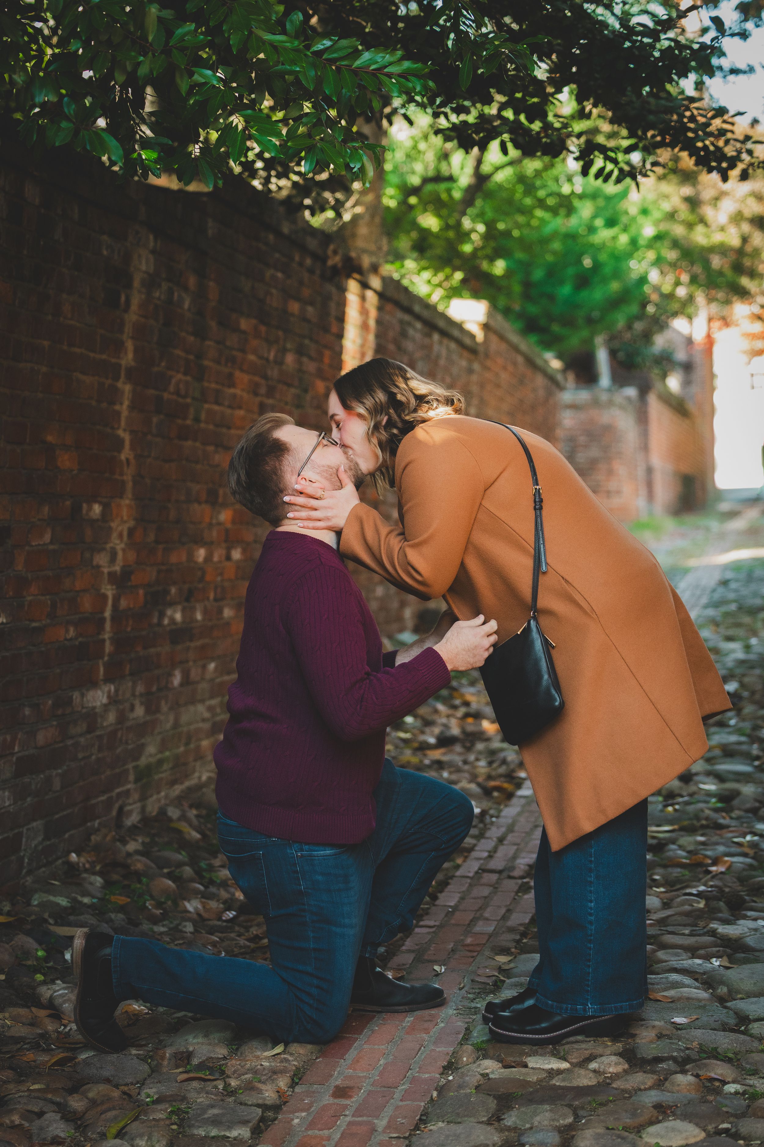 Woman leaning down to kiss her groom on one knee in a sunlit alleyway moments after saying yes