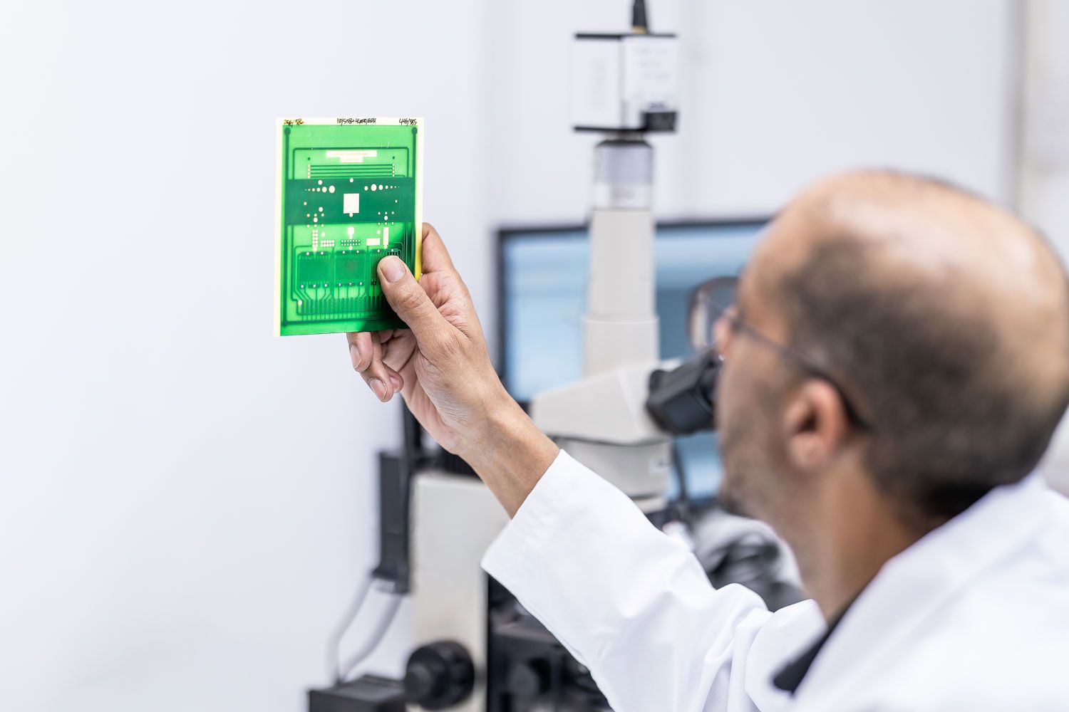 Scientist in white lab coat examining a green circuit board under a microscope in a laboratory setting.