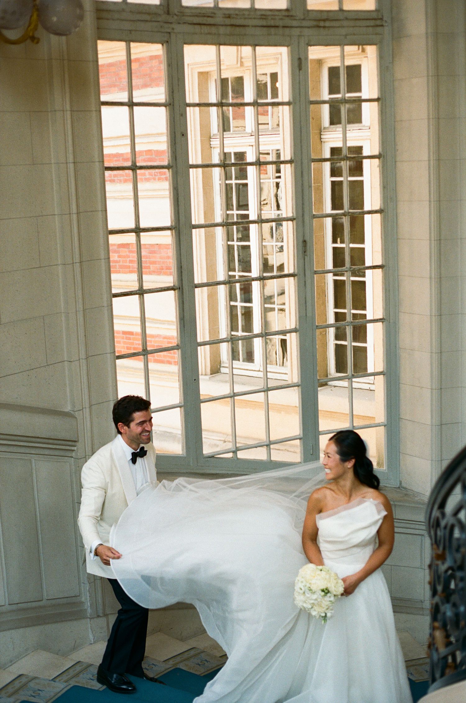 Groom in white tuxedo bride's flowing veil near elegant French windows during Versailles wedding photo session. Versailles film wedding