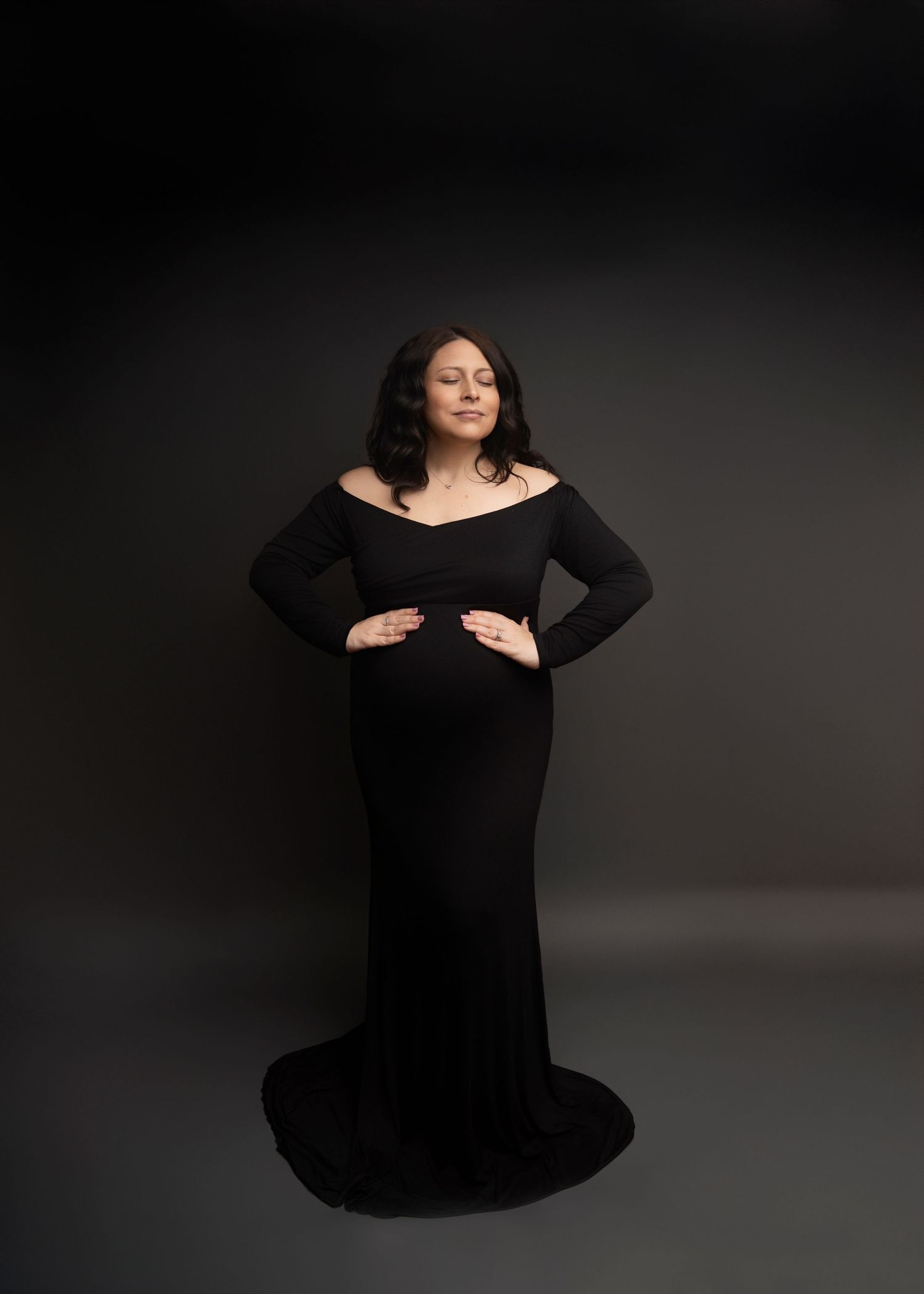 Pregnant woman in elegant black off-shoulder gown poses gracefully against dark studio backdrop.