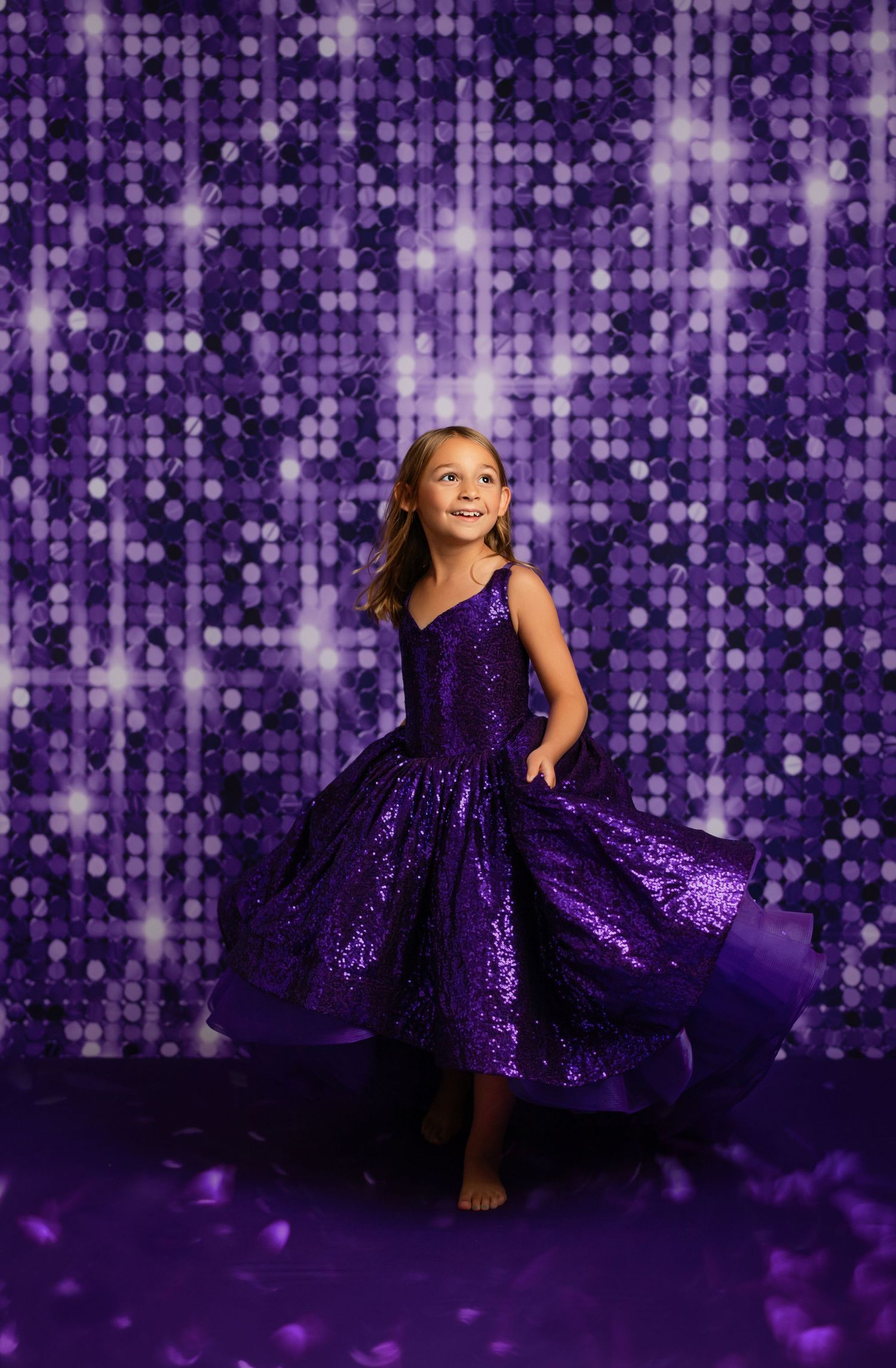 Young girl twirling in a purple sequin dress against a sparkling purple sequin backdrop.