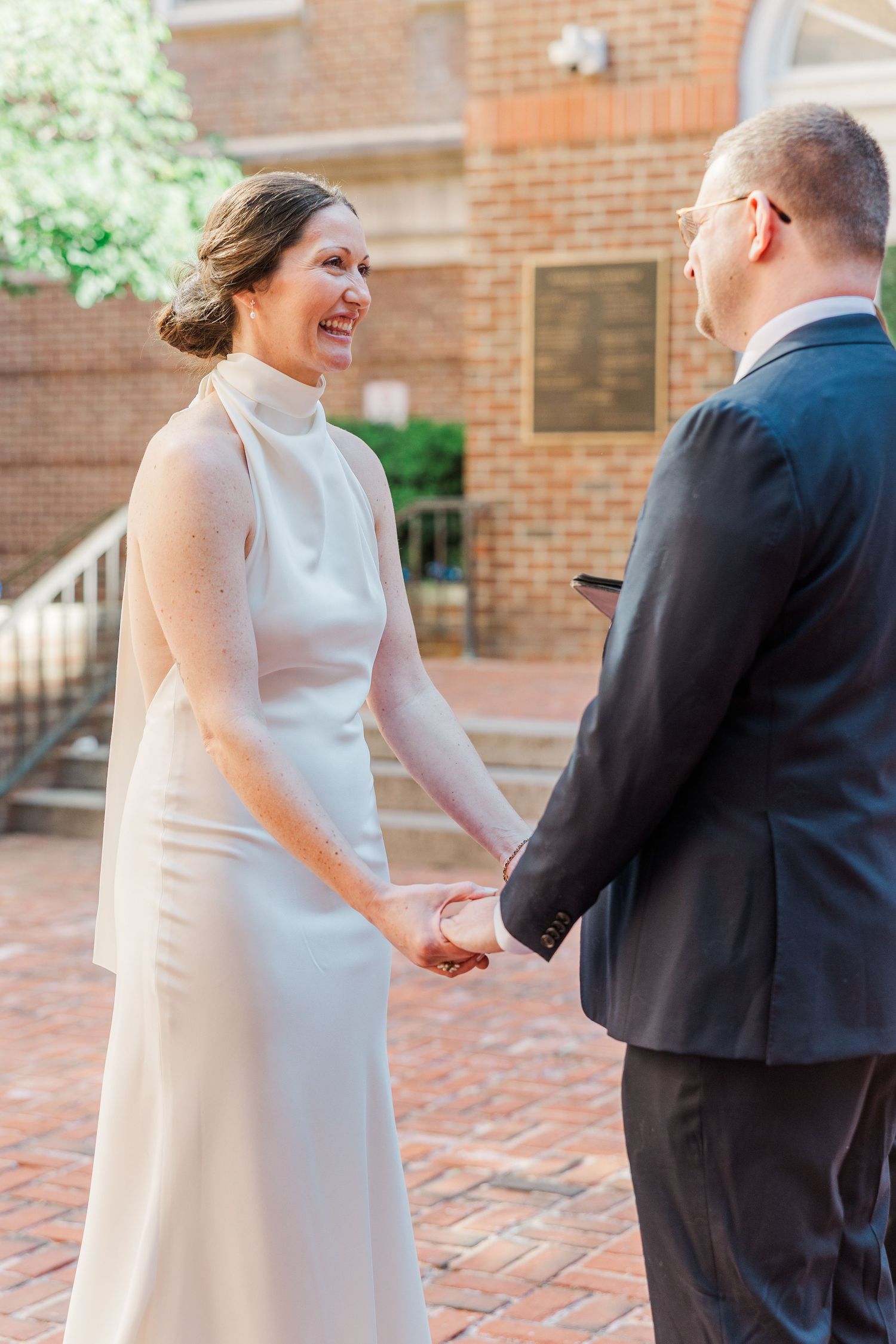 Smiling bride in white gown holds hands with groom outside brick building on wedding day.
