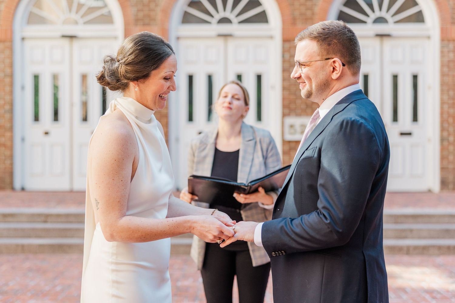 Couple exchanges wedding vows outdoors before officiant at historic brick building ceremony.