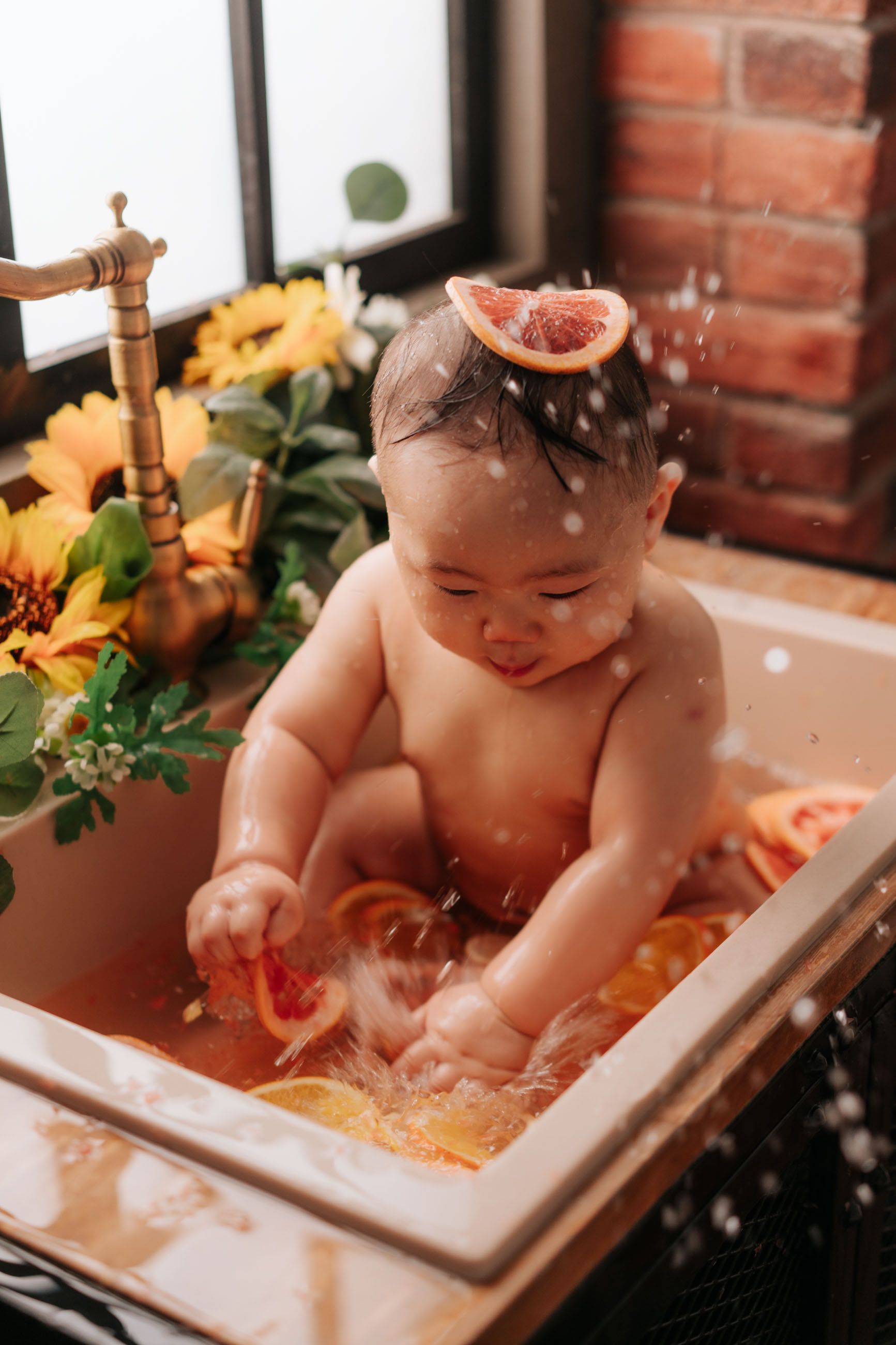 Cute baby sitting in a sink bath with orange slices and a grapefruit slice on head, surrounded by sunflowers.