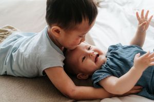 Two Asian toddler siblings lying on a bed, older child kissing baby's cheek while baby smiles and reaches up.
