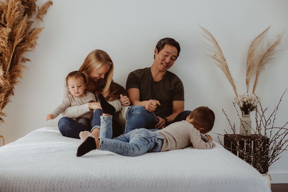 Family of four sitting and playing on a white bed with pampas grass decor in a bright minimalist room.