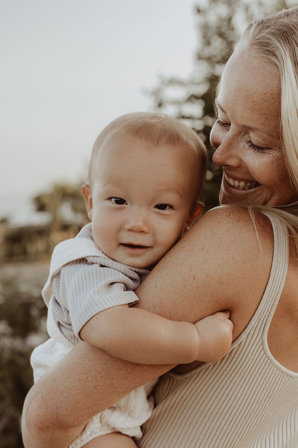 Smiling blonde mother holding her happy baby outdoors in warm natural light.