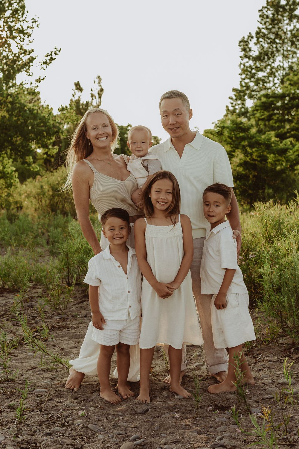 Family of six poses outdoors in neutral whites and beige, parents with four children smiling in a lush green setting.