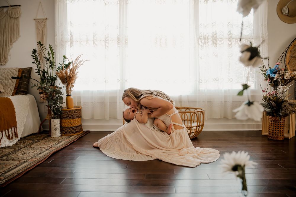 A mother cradles her newborn baby on a wooden floor in a bohemian-styled room with warm natural light.