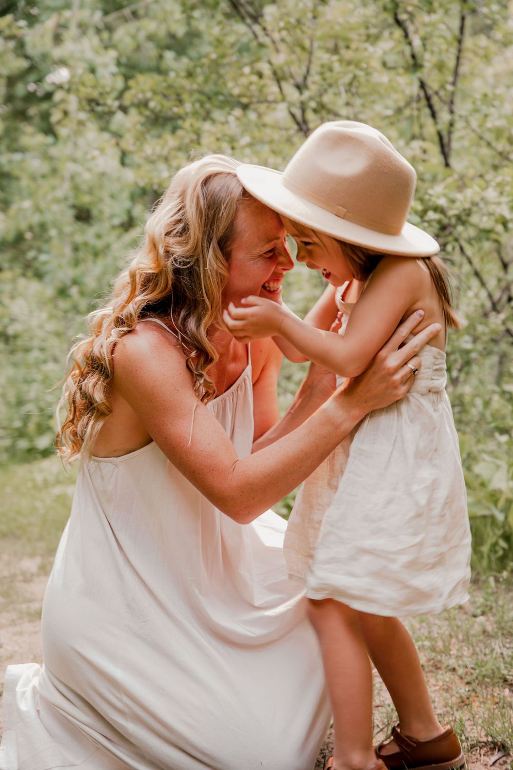 A mother and young daughter share a joyful moment outdoors, dressed in white, with the girl wearing a wide-brim hat.