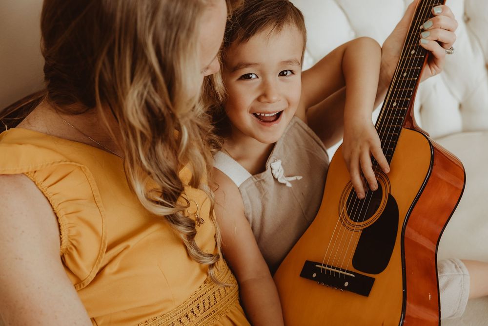 A happy toddler smiles while holding an acoustic guitar with a woman in a yellow dress during a warm family music moment.