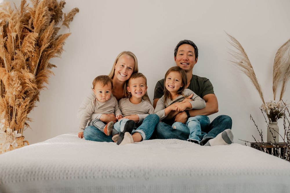 Happy family of five posing together on a white bed with pampas grass decor in a bright, cozy studio setting.