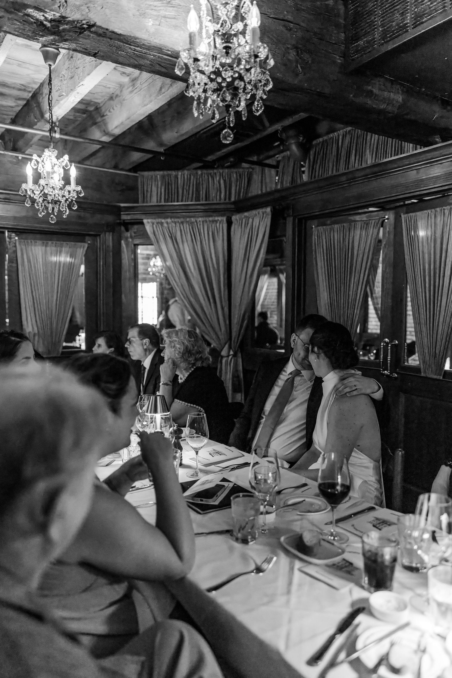 Black and white photo of elegantly dressed guests dining at a formal restaurant with chandeliers and draped curtains.