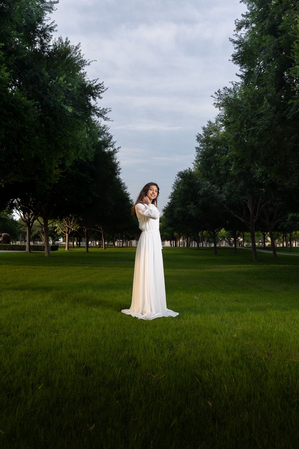 A bride in a flowing white gown stands gracefully on lush green grass surrounded by tall trees in a park.