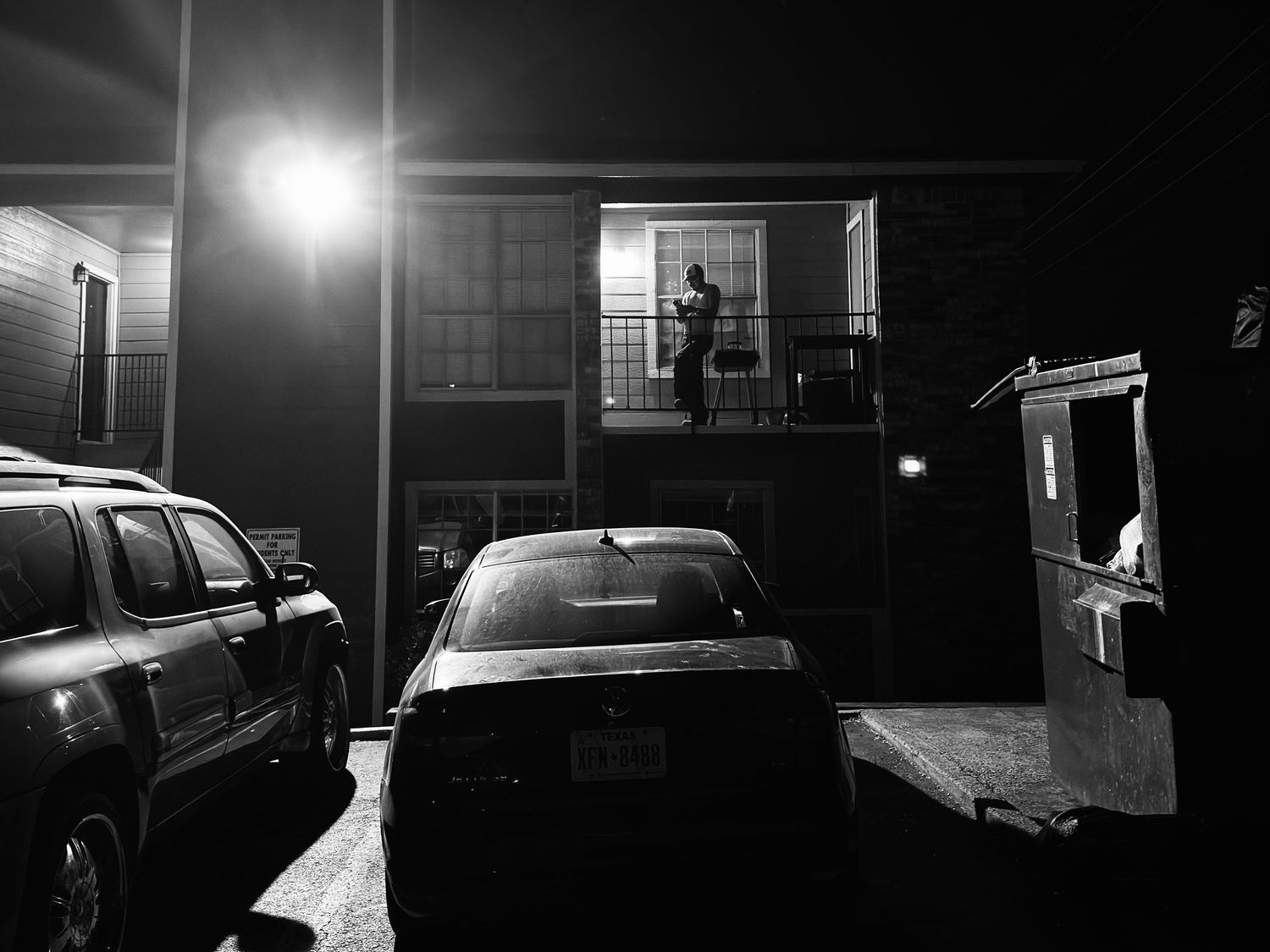 Black and white night photo of a person standing on an apartment balcony, with parked cars below in a dimly lit parking lot.