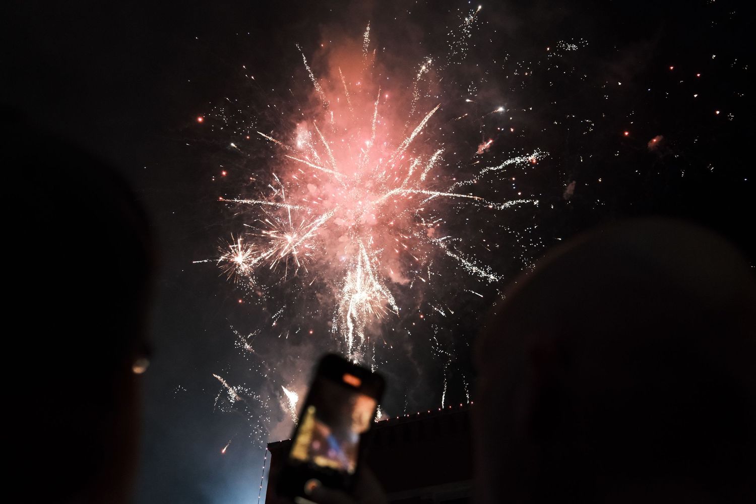 Person capturing colorful fireworks display on smartphone against dark night sky.