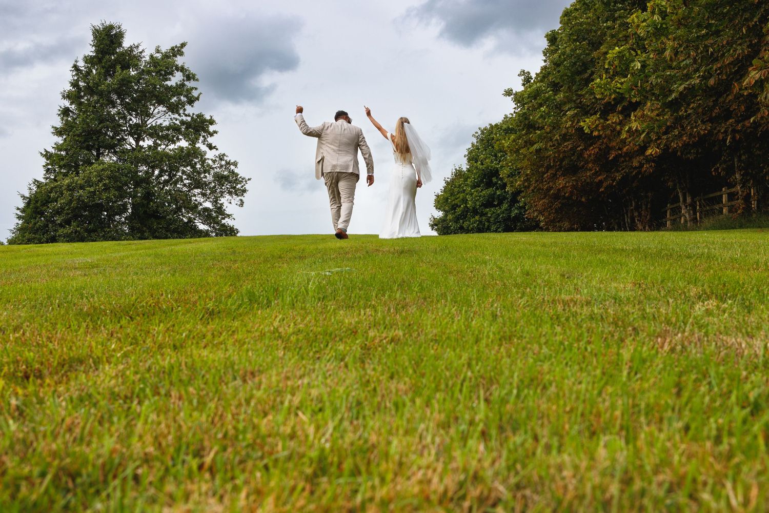 A couple in wedding attire celebrates joyfully at the top of a grassy hill under cloudy skies.