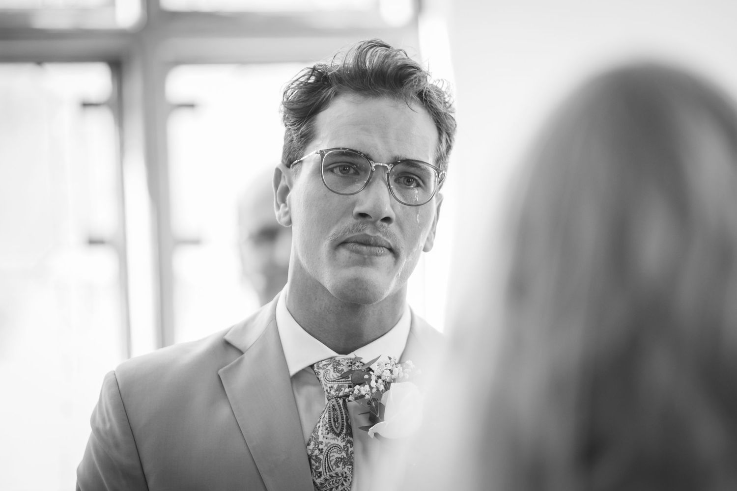 Black and white portrait of a groom in a suit and floral tie wearing glasses at a wedding ceremony.