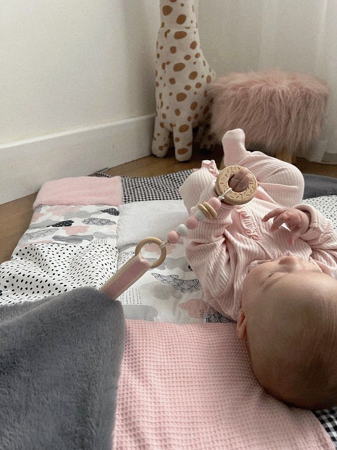 Baby lying on a pink patchwork play mat with a wooden teether ring, surrounded by plush toys and a fluffy pink stool.