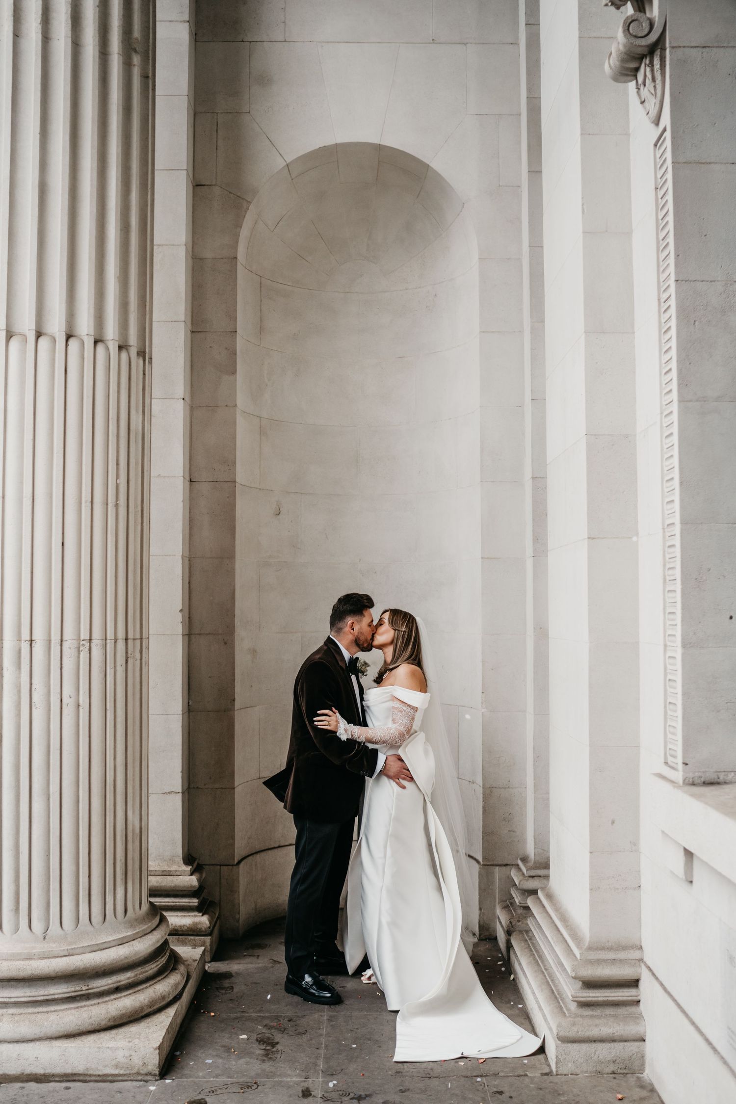A bride and groom share a romantic kiss between grand stone columns at an elegant neoclassical building.