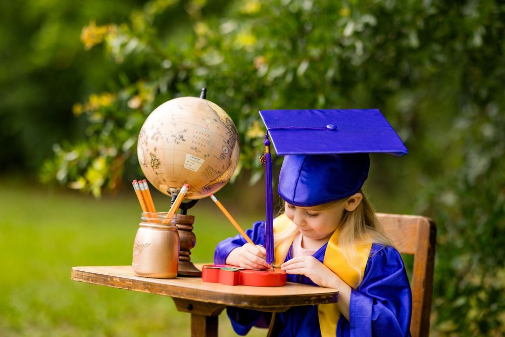 Young girl in blue graduation cap and gown writing at desk with globe outdoors.