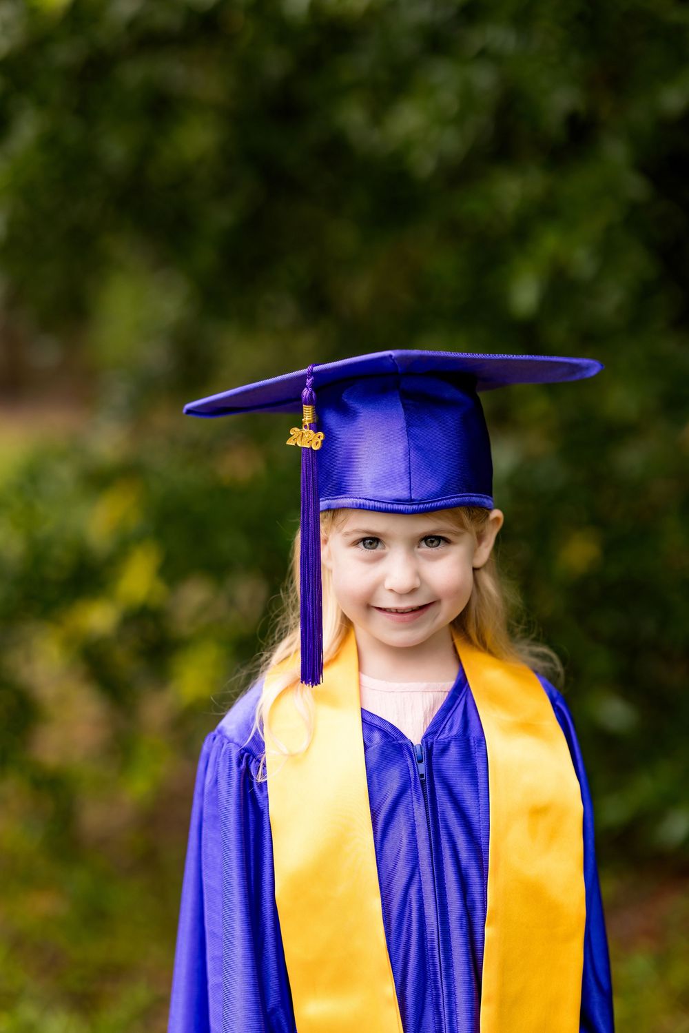 Young girl smiling in blue graduation cap and gown with gold sash, posing outdoors with green trees background.