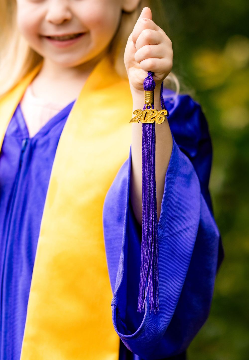 Young girl in blue and gold graduation gown holds up a purple tassel with 2026 charm for kindergarten graduation.