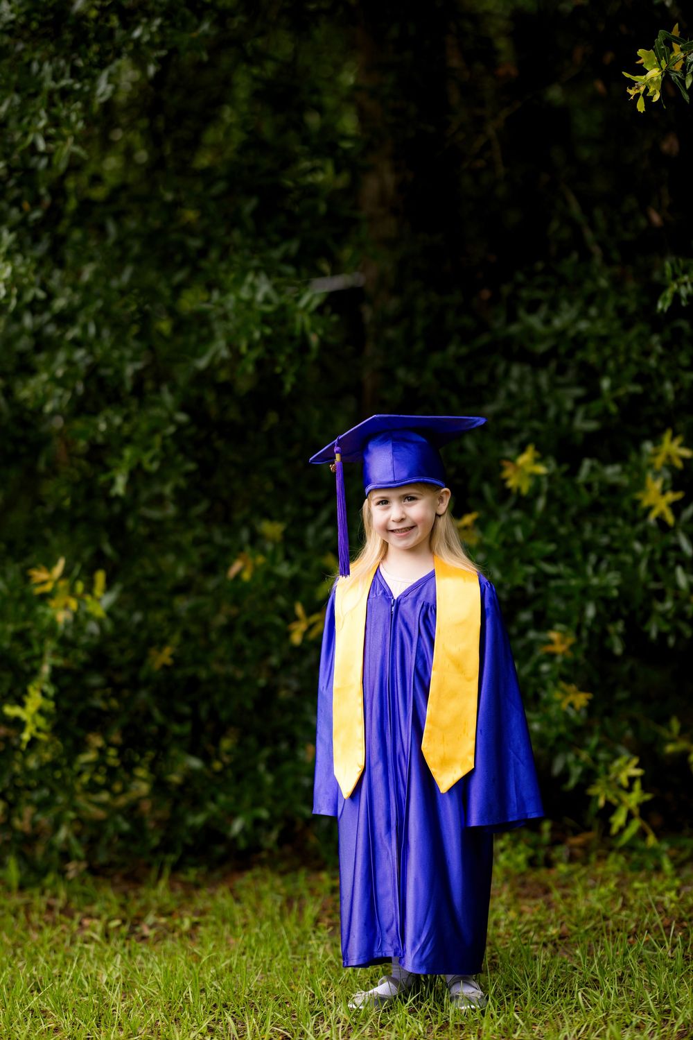 Young girl smiling in blue graduation cap and gown with gold sash, standing outdoors on green grass.
