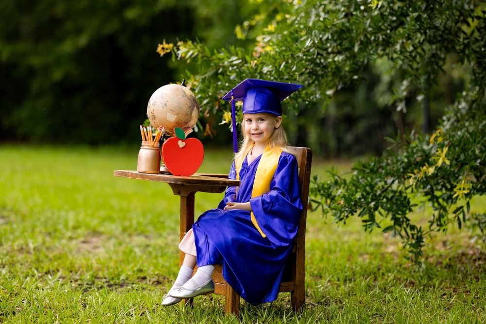 Young girl in blue graduation cap and gown sits outdoors with a globe and red apple, celebrating kindergarten graduation.