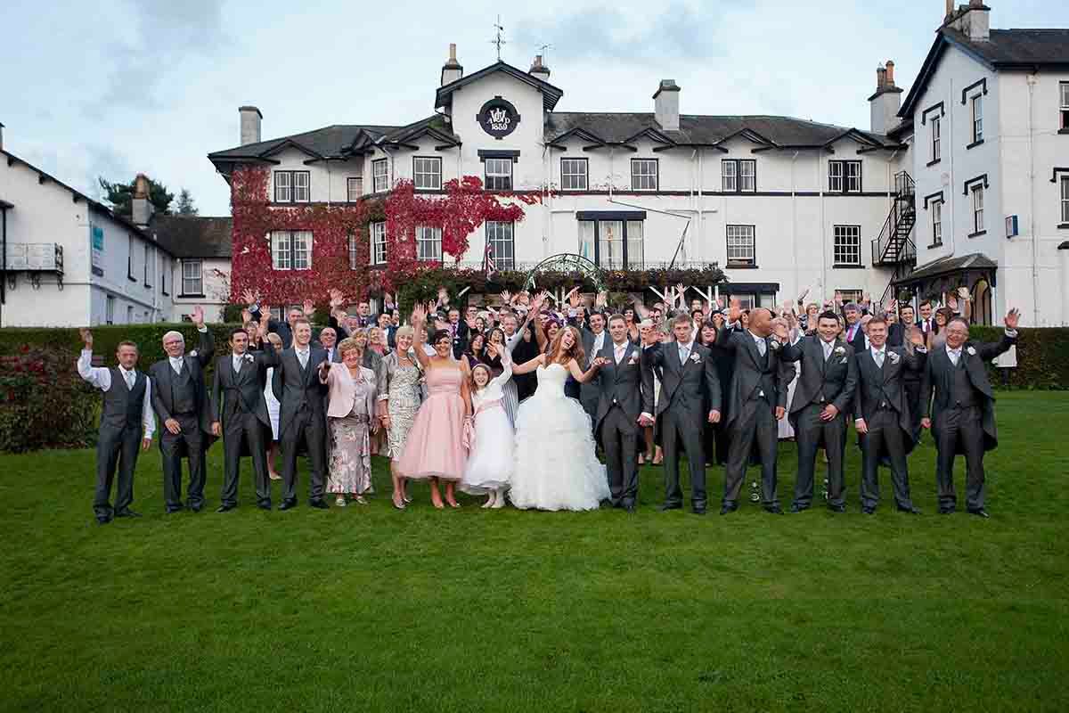 Large wedding party celebrating outdoors in front of a grand manor house, bride and groom surrounded by guests cheering.