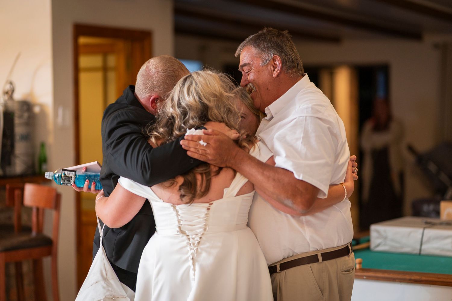 Bride and groom share an emotional group hug with family members during a heartfelt wedding celebration indoors.