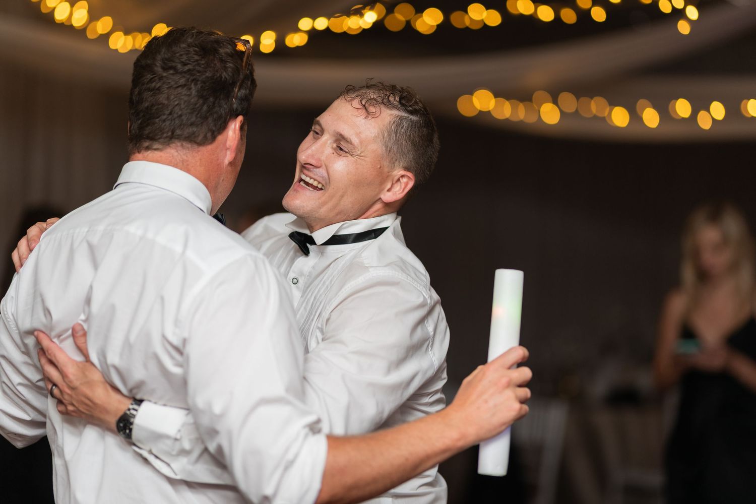 Two men in white dress shirts dancing and laughing together at a wedding reception with bokeh lights in background.