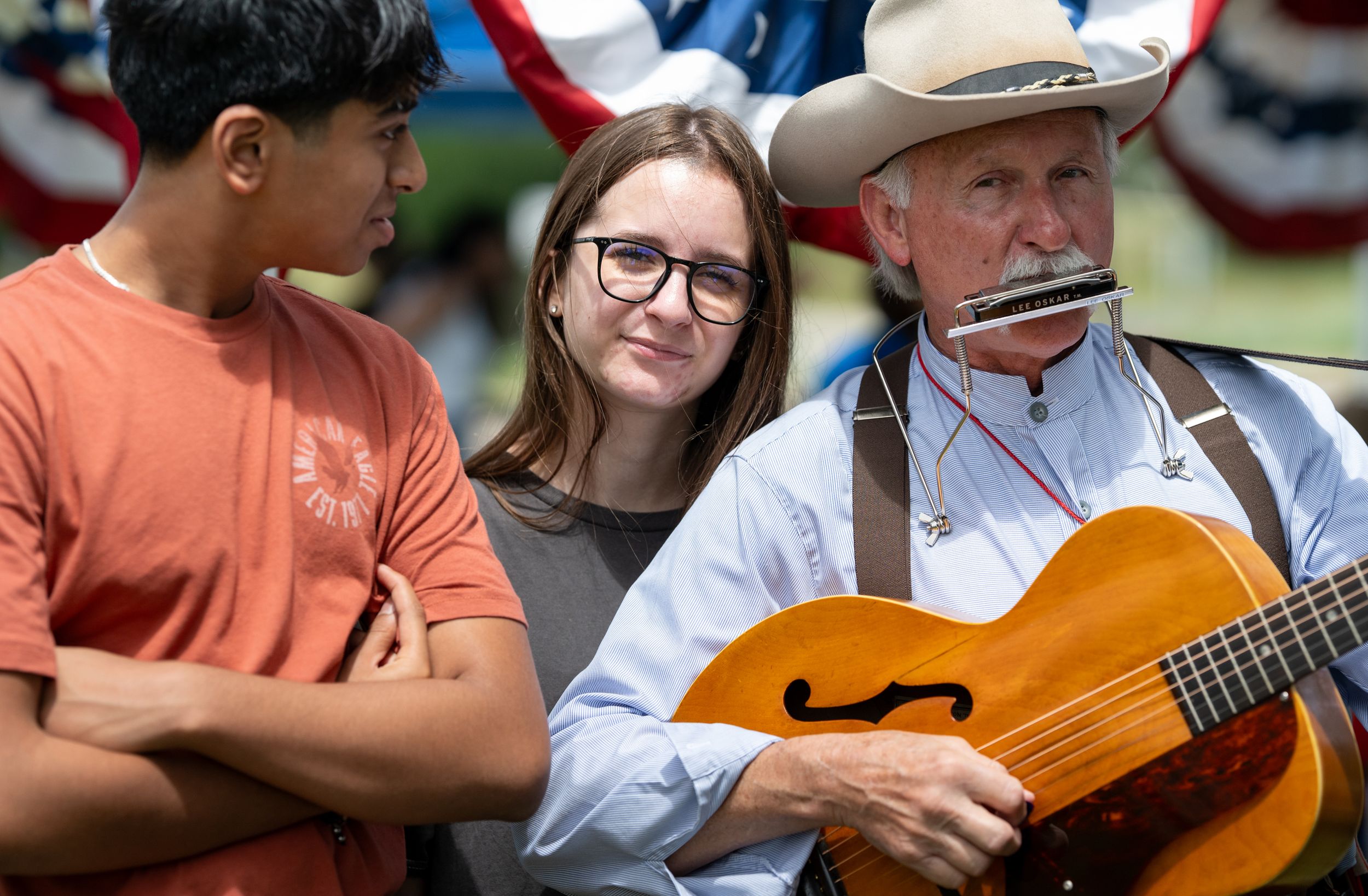 Cowboy musician plays harmonica and guitar while two young people watch at an outdoor patriotic event.